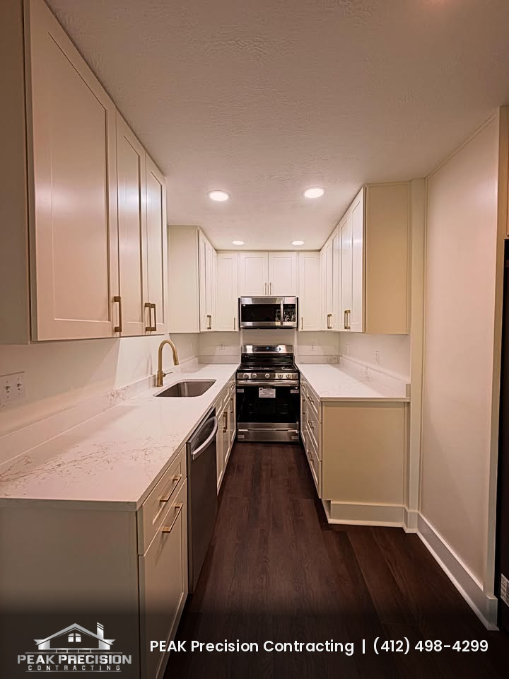 Renovated galley kitchen with white shaker cabinets, light quartz-style countertops, stainless range and microwave, recessed lighting, and dark wood-look flooring by Peak Precision Contracting